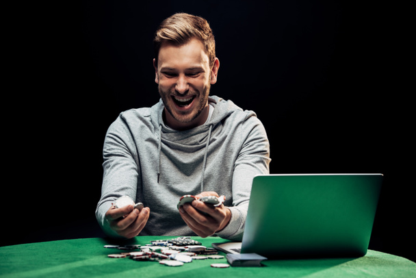 szczęśliwy człowiek trzyma żetony pokera w pobliżu laptopa na stole pokerowym na białym tle  - Zdjęcie, obraz