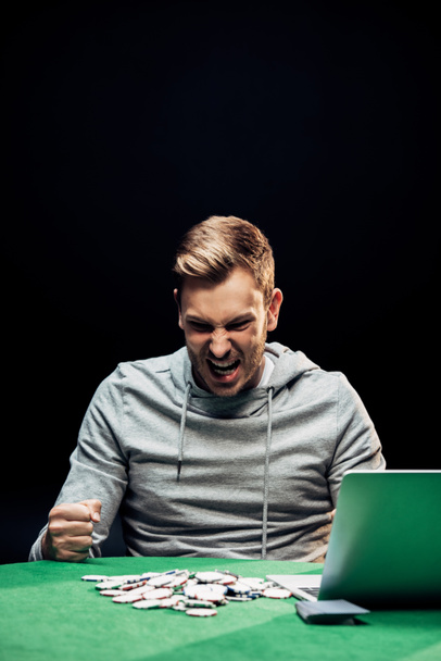 angry man with clenched fists near laptop on poker table isolated on black  - Photo, Image