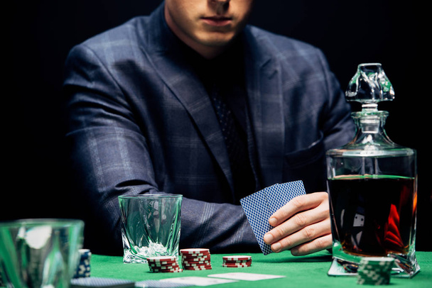 KYIV, UKRAINE - AUGUST 20, 2019: cropped view of man holding playing cards near poker chips isolated on black  - Photo, image