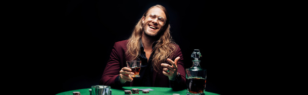 panoramische shot van gelukkige bebaarde man in oog glazen Holding glas met whisky in de buurt van speelkaarten geïsoleerd op zwart  - Foto, afbeelding
