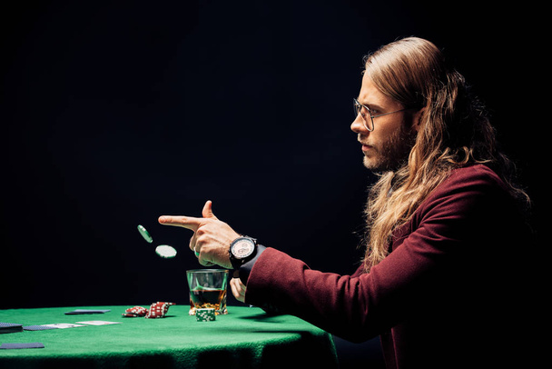 Seitenansicht des Mannes mit Brille in der Nähe von Glas mit Whiskey werfen in Luft Pokerchips isoliert auf schwarz  - Foto, Bild
