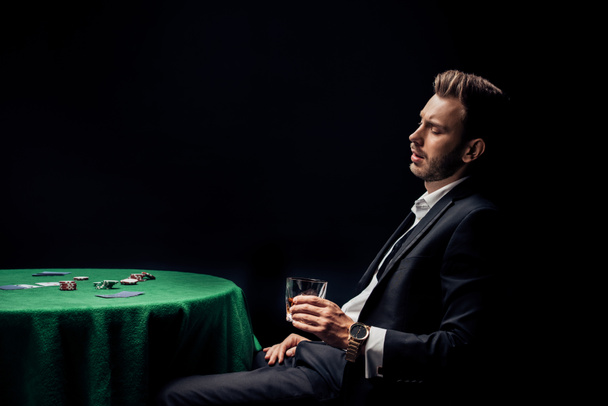 Verärgerter bärtiger Mann hält Glas in der Nähe von Pokertisch isoliert auf schwarz  - Foto, Bild