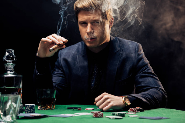 schöner Mann raucht Zigarre in der Nähe des Pokertisches auf schwarz mit Rauch  - Foto, Bild