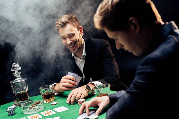 KYIV, UKRAINE - AUGUST 20, 2019: selective focus of cheerful man playing poker near friend and poker chips on black with smoke  - Foto, imagen