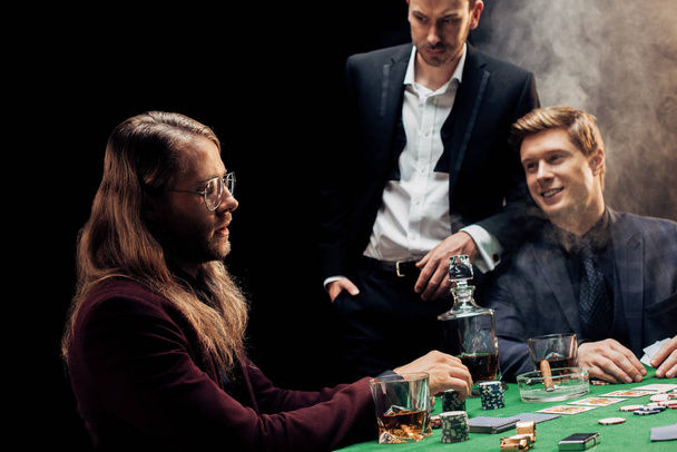 KYIV, UKRAINE - AUGUST 20, 2019: handsome friends gesturing while playing poker on black with smoke  - Fotografie, Obrázek