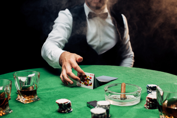 KYIV, UKRAINE - AUGUST 20, 2019: cropped view of croupier touching playing cards near poker table on black with smoke  - Valokuva, kuva