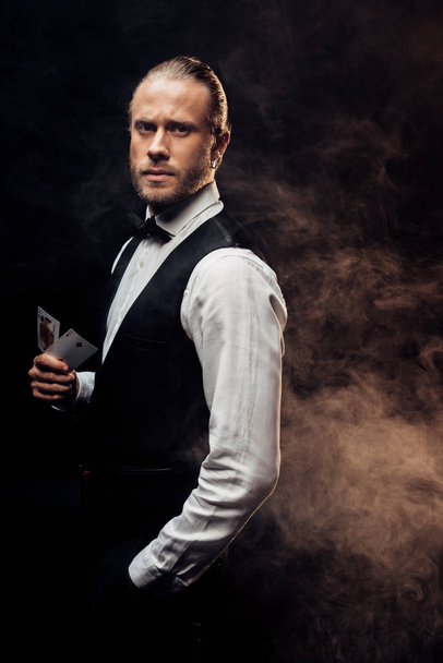 KYIV, UKRAINE - AUGUST 20, 2019: handsome croupier holding playing cards on black with smoke - Foto, imagen
