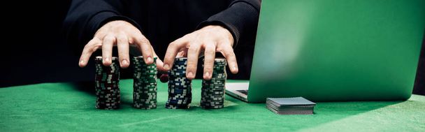 KYIV, UCRANIA - 20 de agosto de 2019: plano panorámico del hombre tocando fichas de póquer cerca del ordenador portátil aislado en negro
 - Foto, Imagen