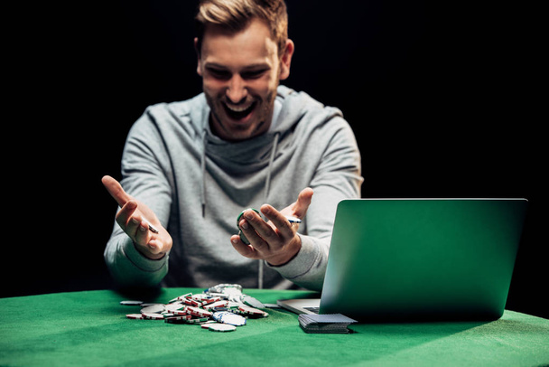 KYIV, UKRAINE - AUGUST 20, 2019: selective focus of happy man looking at poker chips near laptop isolated on black  - Photo, Image