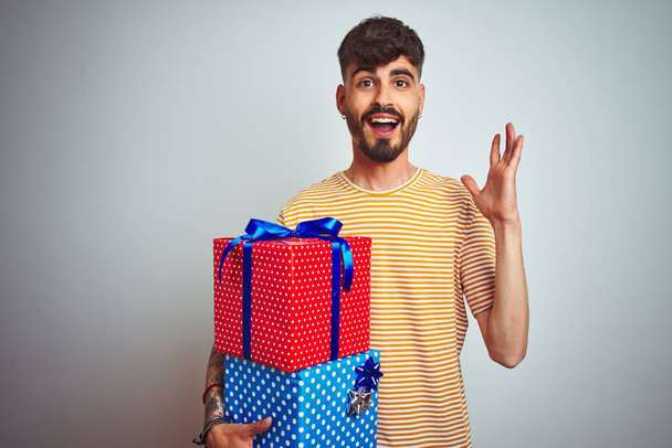 Doğum günü hediyeleri taşıyan dövmeli genç adam izole edilmiş beyaz arka planda mutlu ve heyecanlı duruyor, kazanan yüz ifadesi zaferi büyük bir gülümsemeyle kutluyor ve ellerini kaldırıyor. - Fotoğraf, Görsel