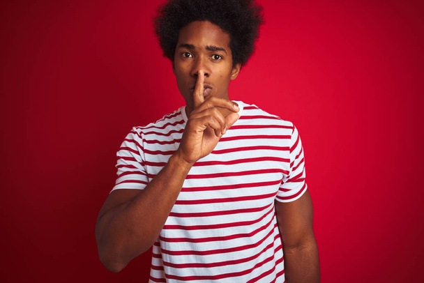 junger afrikanisch-amerikanischer Mann mit Afro-Haaren, der gestreiftes T-Shirt über isoliertem rotem Hintergrund trägt und mit dem Finger auf den Lippen um Ruhe bittet. Schweigen und geheimes Konzept. - Foto, Bild