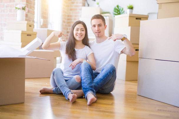 Jeune beau couple assis sur le sol à la nouvelle maison autour de boîtes en carton avec visage surprise pointant vers lui-même
 - Photo, image