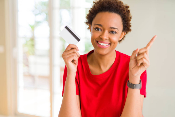 Young african american woman holding credit card very happy pointing with hand and finger to the side - Фото, изображение