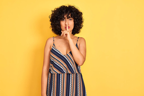 Arab woman with curly hair wearing striped colorful dress over isolated yellow background asking to be quiet with finger on lips. Silence and secret concept. - Valokuva, kuva
