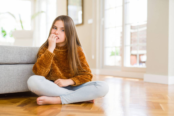Belle jeune fille enfant assis sur le sol à la maison regardant stressé et nerveux avec les mains sur la bouche mordant ongles. Problème d'anxiété
. - Photo, image
