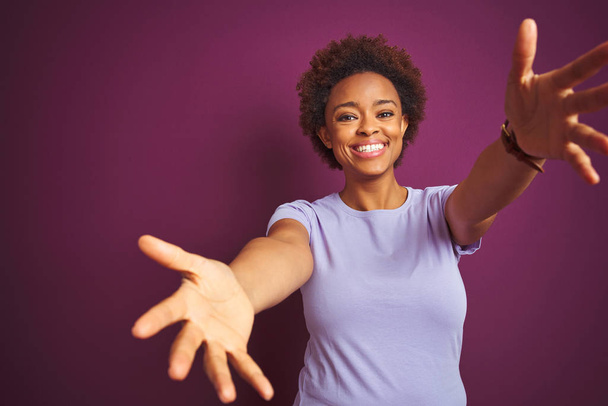 Giovane bella donna afro americana con i capelli afro su sfondo viola isolato guardando la fotocamera sorridente a braccia aperte per l'abbraccio. Espressione allegra che abbraccia la felicità
. - Foto, immagini