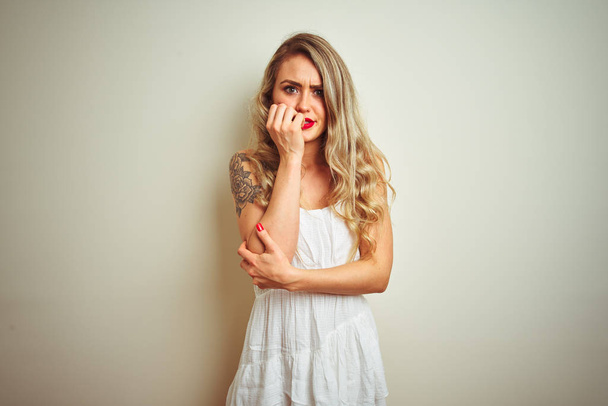 junge schöne Frau in lässigem Kleid, die vor weißem isolierten Hintergrund steht, sieht gestresst und nervös aus, die Hände auf den Mund beißenden Nägeln. Angstproblem. - Foto, Bild