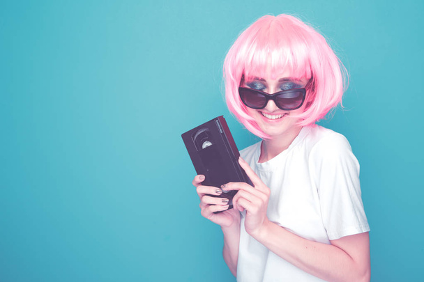 Концепция стиля 90-х. Молодая женщина с розовым градом, держащая стаканы и кассету, стоящую в голубой студии.
. - Фото, изображение