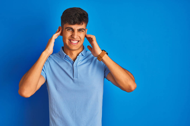 隔絶された青い背景の上にカジュアルなポロシャツを着た若いインド人男性は、大きな音楽のノイズに悩まされた表情で耳を覆います。ろう者の概念. - 写真・画像