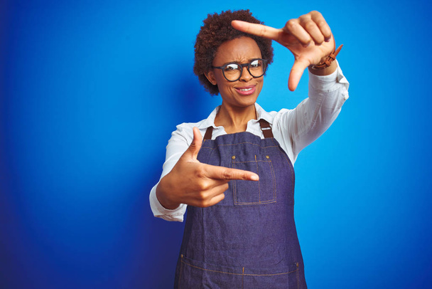 Молода афроамериканська жінка, власник магазину, одягнена в діловий фартух над синім фоном, посміхається, роблячи рамку руками і пальцями з щасливим обличчям. Творчість і фотографія. - Фото, зображення