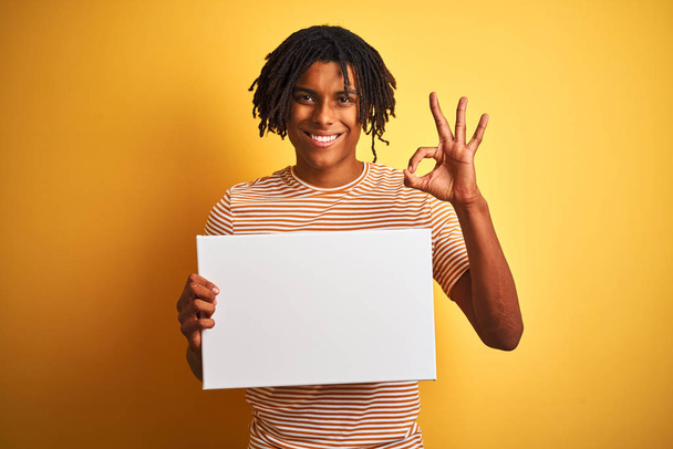 Afroamerikanischer Mann mit Dreadlocks, die Banner über isoliertem gelbem Hintergrund halten, macht ok Zeichen mit Fingern, ausgezeichnetes Symbol - Foto, Bild
