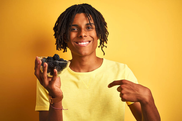 Afro-Amerikaanse met dreadlocks houden bramen over geïsoleerde gele achtergrond met verrassing gezicht wijzend vinger naar zichzelf - Foto, afbeelding
