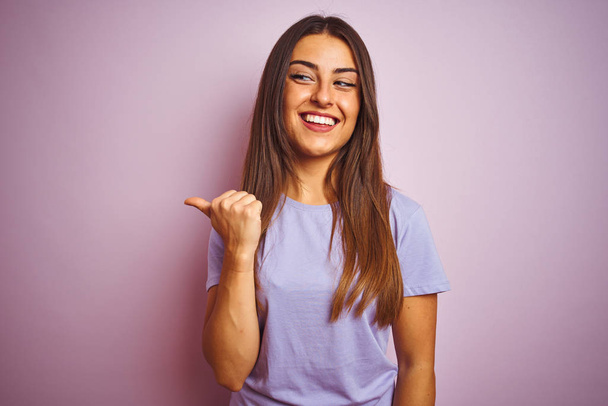 Young beautiful woman wearing casual t-shirt standing over isolated pink background smiling with happy face looking and pointing to the side with thumb up. - Photo, Image