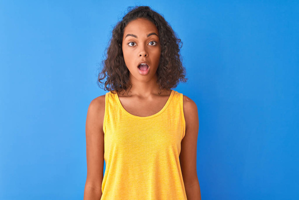 Young brazilian woman wearing yellow t-shirt standing over isolated blue background afraid and shocked with surprise expression, fear and excited face. - Fotoğraf, Görsel