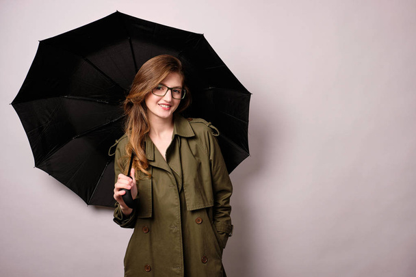 A blonde girl in glasses and a raincoat is standing with a black umbrella and smiling at the camera with her hand in her pocket - Fotografie, Obrázek