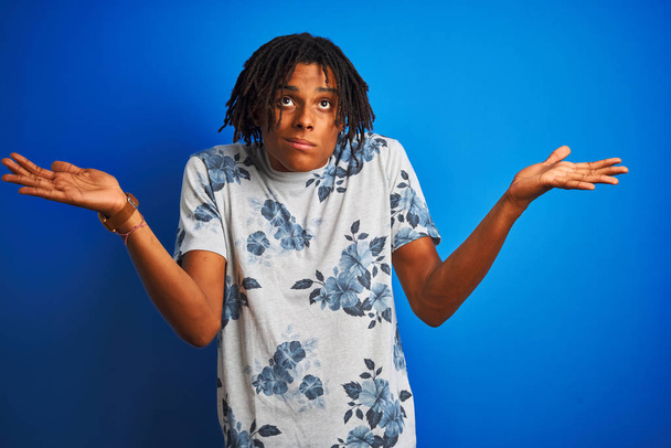 Afro-Amerikaanse man met dreadlocks dragen zomer bloemen t-shirt over geïsoleerde blauwe achtergrond clueless en verwarde uitdrukking met armen en handen opgeheven. Twijfelachtig concept. - Foto, afbeelding