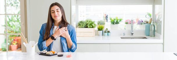 Wide angle picture of beautiful young woman eating delivery sushi smiling with hands on chest with closed eyes and grateful gesture on face. Health concept. - Φωτογραφία, εικόνα
