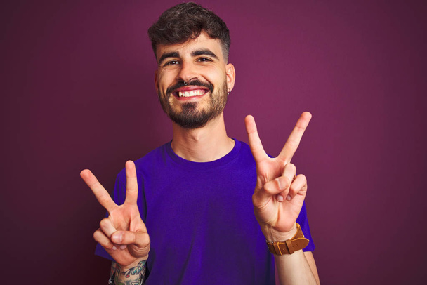 Młody człowiek z tatuażem w koszulce stojący nad odizolowanym fioletowym tłem uśmiechnięty do kamery pokazujący palce robiące znak zwycięstwa. Numer dwa.. - Zdjęcie, obraz