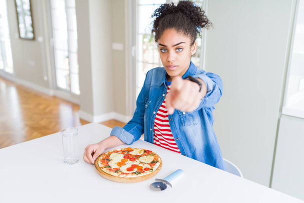 Joven mujer afroamericana comiendo pizza casera de queso mozzarella señalando con el dedo a la cámara y a usted, signo de mano, gesto positivo y seguro de la parte delantera
 - Foto, Imagen