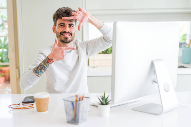 Bilgisayarda çalışan genç adam gülümsüyor ve mutlu yüzlü eller ve parmaklarla çerçeve yapıyor. Yaratıcılık ve fotoğrafçılık kavramı. - Fotoğraf, Görsel