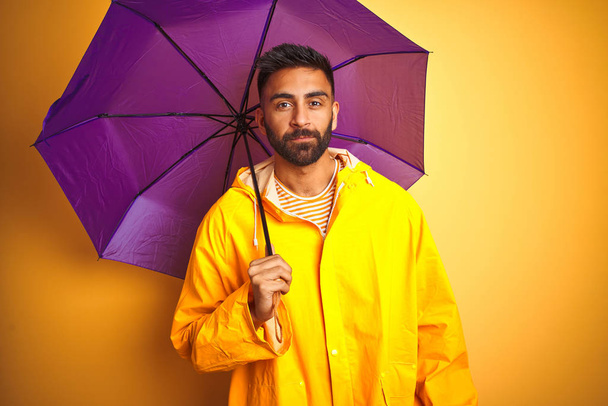 Young indian man wearing raincoat and purple umbrella over isolated yellow background with a confident expression on smart face thinking serious - Fotografie, Obrázek