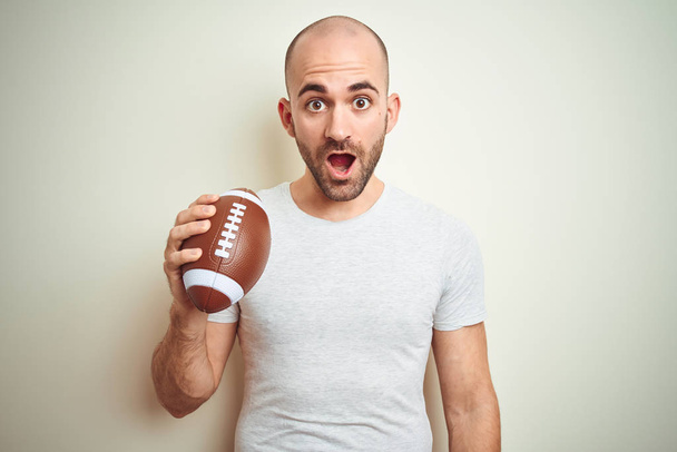 Fiatal férfi tartja rögbi amerikai futball labda felett elszigetelt háttér ijedt sokk egy meglepetés arc, fél, és izgatott a félelem kifejezés - Fotó, kép