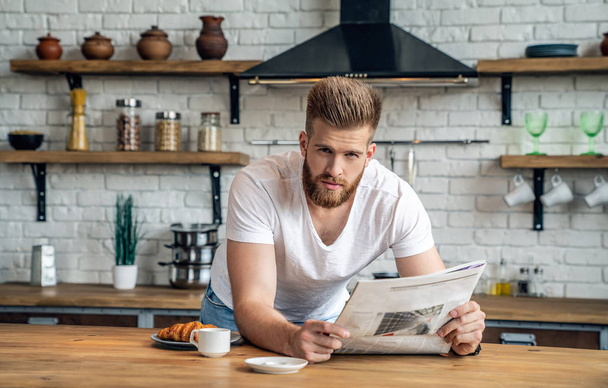 Początek dnia z dobrej nowiny. Przystojny młody człowiek czytania gazety poważnie podczas picia kawy i śniadanie w kuchni, i looning do kamery - Zdjęcie, obraz