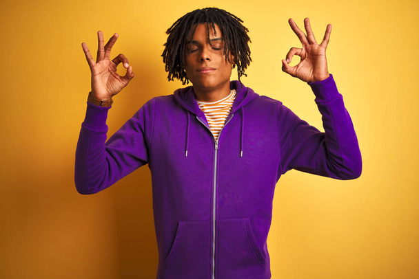 Африканський американець з дредами в пурпуровому светрі через ізольований фіолетовий фон відпочиває і посміхається закритими очима, роблячи жест медитації пальцями. Поняття йоги. - Фото, зображення