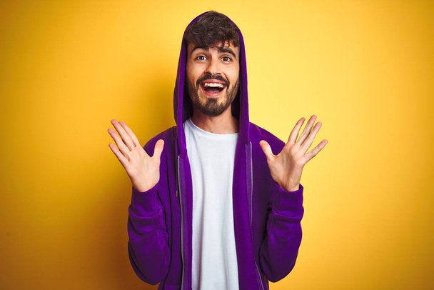 Jeune homme avec tatouage portant sweat-shirt violet avec capuche sur fond jaune isolé célébrant fou et étonné pour le succès avec les bras levés et les yeux ouverts hurlant excités. Concept gagnant
 - Photo, image