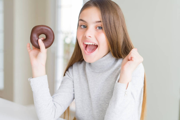 Piękna młoda dziewczyna w czekoladzie jedząc pączka krzycząc dumny i świętując zwycięstwo i sukces bardzo podekscytowany, radosne emocje - Zdjęcie, obraz