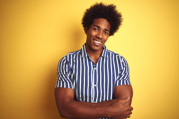 Amerikai férfi afro haj visel csíkos ing felett áll elszigetelt sárga háttér boldog arc mosolyogva keresztbe karok nézi a kamerát. Pozitív személy. - Fotó, kép