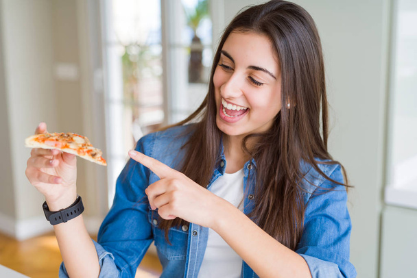 Όμορφη νεαρή γυναίκα τρώει μια φέτα νόστιμη πίτσα πολύ χαρούμενος δείχνοντας με το χέρι και το δάχτυλο - Φωτογραφία, εικόνα