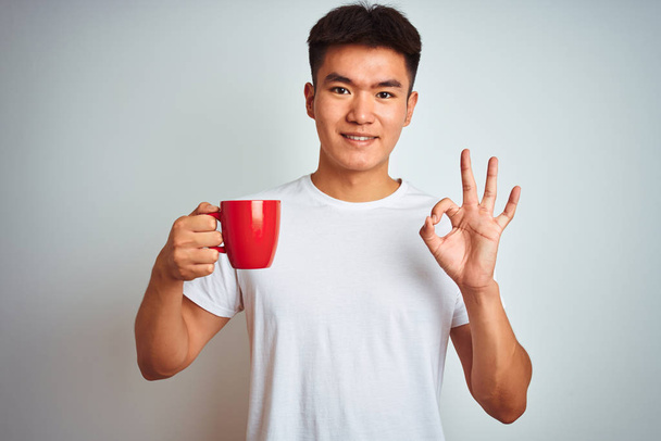 Asian chinese man holding red cup of coffee standing over isolated white background doing ok sign with fingers, excellent symbol - Foto, Imagem