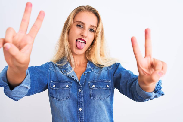 Young beautiful woman wearing casual denim shirt standing over isolated white background smiling with tongue out showing fingers of both hands doing victory sign. Number two. - Photo, Image