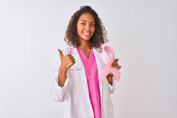 junge brasilianische Ärztin hält Krebsband über isoliertem weißem Hintergrund glücklich mit großem Lächeln tun ok Zeichen, Daumen nach oben mit den Fingern, ausgezeichnetes Zeichen - Foto, Bild