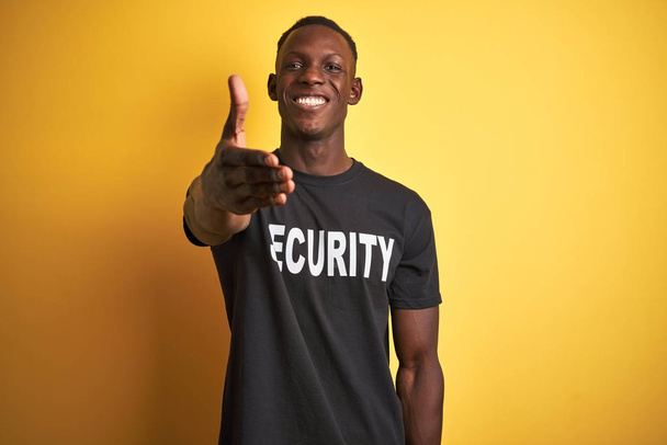 Afroameričan ochranka muž v bezpečnostní uniformě přes izolované žluté pozadí s úsměvem přátelské nabízí potřesení rukou jako pozdrav a přivítání. Úspěšná práce. - Fotografie, Obrázek