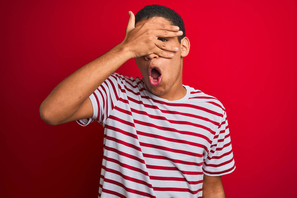 Jonge knappe arabische man draagt gestreept t-shirt over geïsoleerde rode achtergrond gluren in shock bedekken gezicht en ogen met de hand, kijken door vingers met beschaamde uitdrukking. - Foto, afbeelding