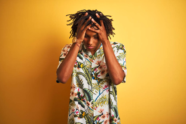Афро-человек с дредами на отдыхе в летней рубашке на изолированном желтом фоне страдает от головной боли отчаянно и стресс, потому что боль и мигрень. Руки на голову
. - Фото, изображение