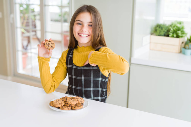 Hermosa niña comiendo galletas de chispas de chocolate con la cara sorpresa señalando el dedo a sí mismo
 - Foto, Imagen