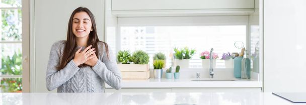 Wide angle picture of beautiful young woman sitting on white table at home smiling with hands on chest with closed eyes and grateful gesture on face. Health concept. - Foto, afbeelding